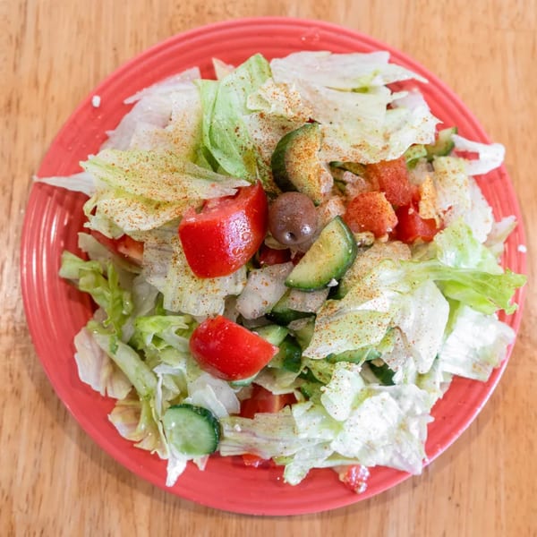 A vibrant salad with lettuce, tomatoes, cucumbers, and olives, sprinkled with seasoning on a red plate.