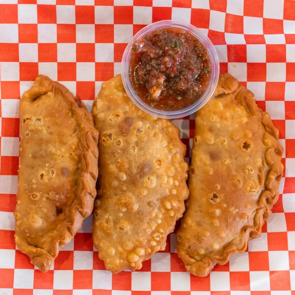 Three golden-brown fried empanadas with a side of dark red dipping sauce on red and white checkered paper.