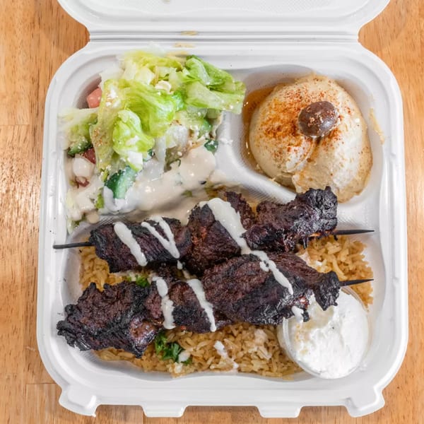 Takeout container with two beef shish kebabs, rice, salad, hummus, and a side of white sauce.
