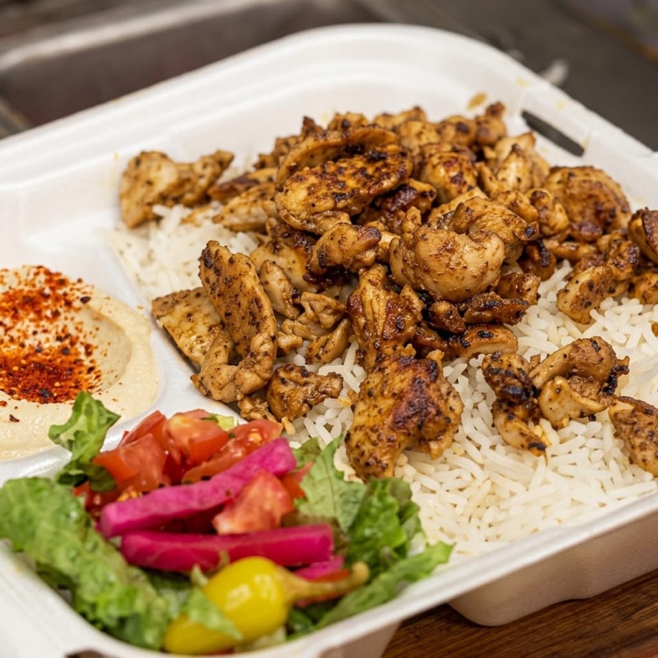 Grilled chicken and mushrooms over white rice with hummus, salad, pickled vegetables, and lemon on a platter.