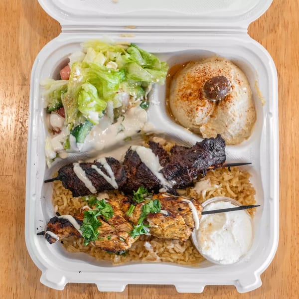 Takeout container with two skewers of grilled meat, rice, salad, hummus, and a side of sauce.