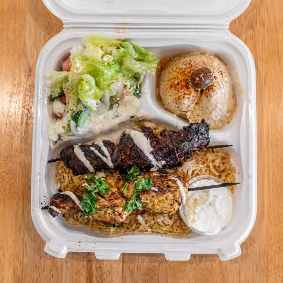 Open takeout container filled with grilled meats, rice, salad, pita, sauces, and a falafel-like side dish.