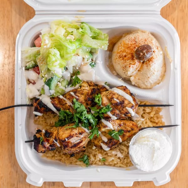 Grilled chicken skewers with rice, salad, hummus, and a side of white sauce in a takeout container.