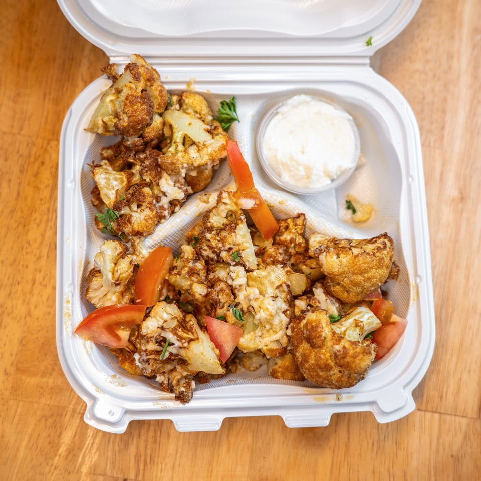 Open takeout container filled with seasoned roasted cauliflower, tomato wedges, herbs, and a cup of creamy dipping sauce.