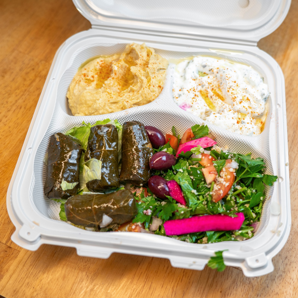 Open takeout container filled with hummus, creamy dip, stuffed grape leaves, olives, pickled vegetables, and fresh herb salad.