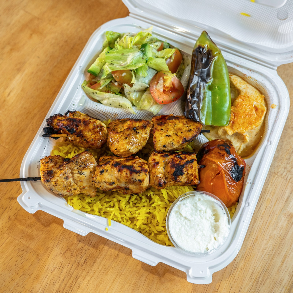 Grilled chicken skewers with yellow rice, salad, roasted peppers, hummus, and yogurt sauce in a takeout container.