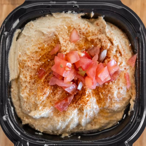 Creamy hummus topped with diced tomatoes and paprika in a black takeout container.