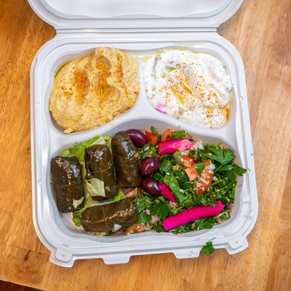 Open takeout container holding hummus, creamy dip, stuffed grape leaves, tabbouleh, olives, and pickled turnips.
