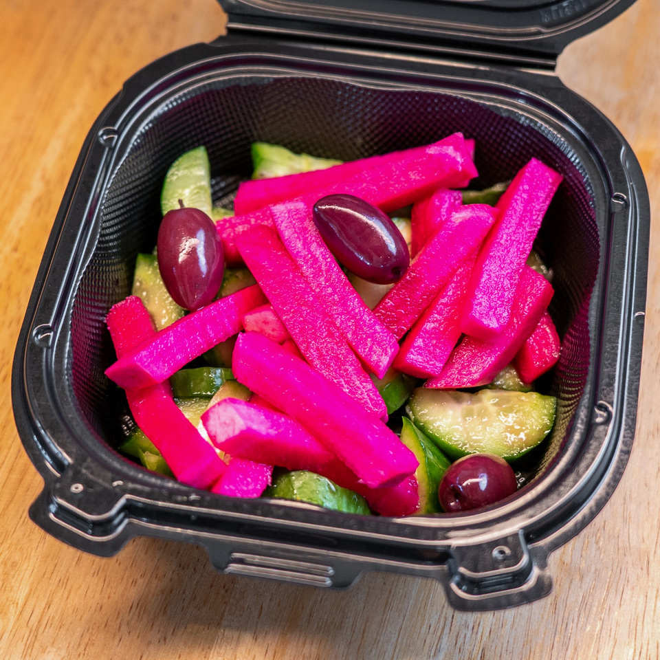Open black container filled with bright pink pickled vegetables, cucumber slices, green peppers, and dark olives on a wooden table.