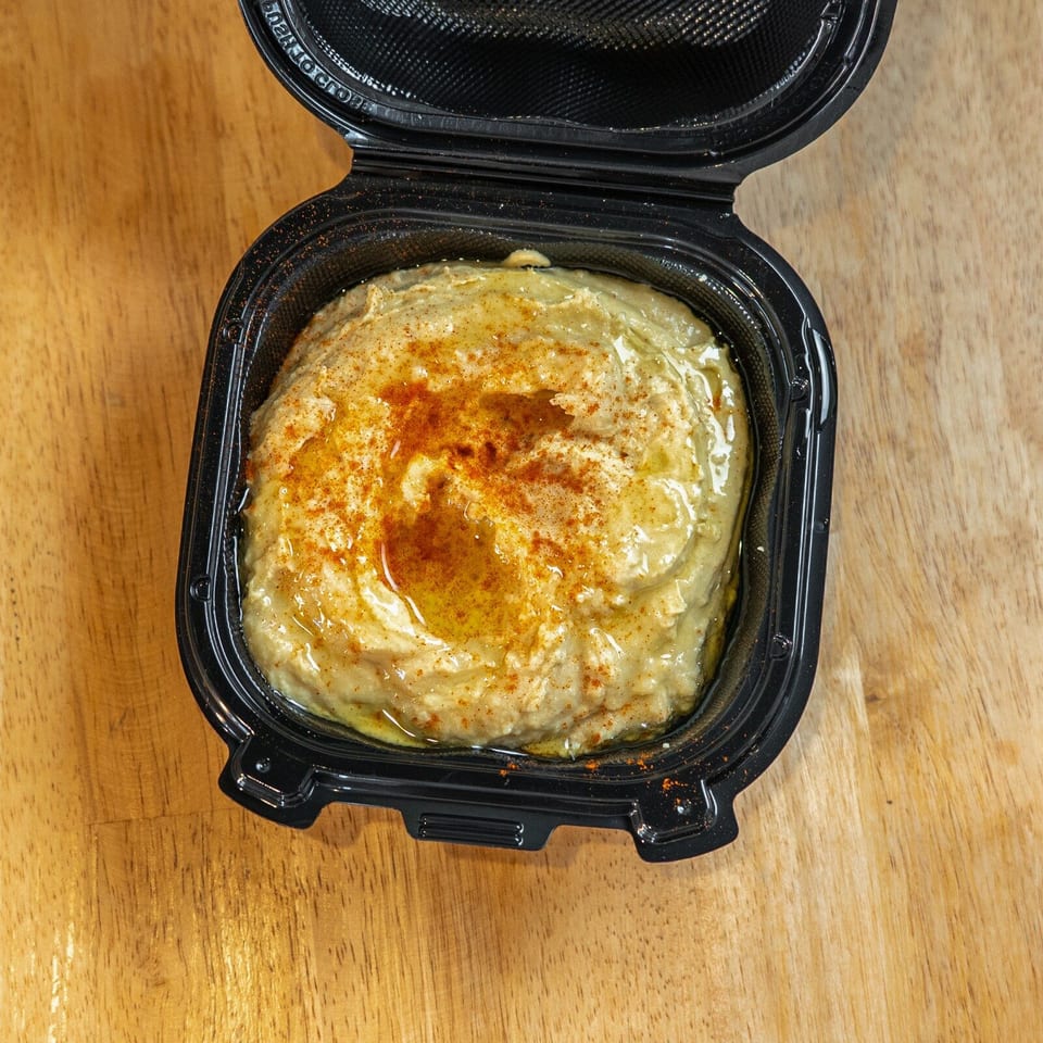 Open black takeout container holding creamy hummus topped with olive oil and paprika on a wooden table.