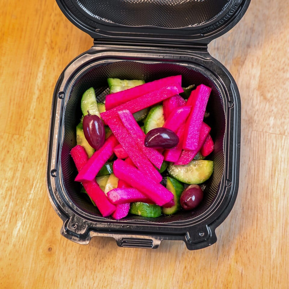 Open takeout container holding bright pink pickled vegetables, cucumber pieces, and dark olives on a wooden table.