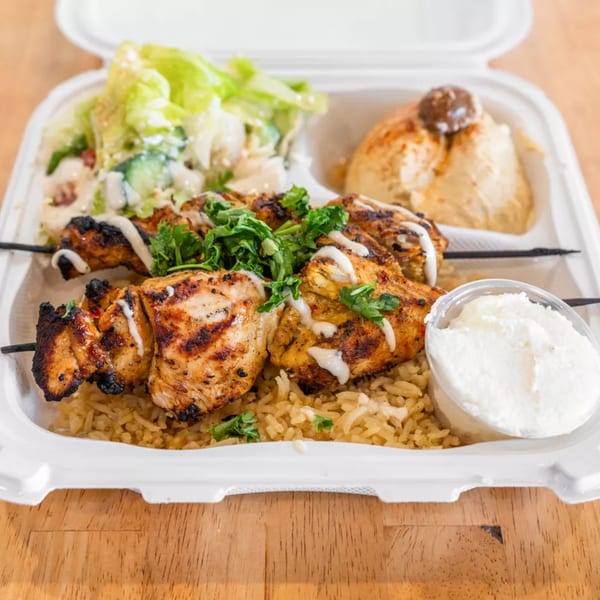 Grilled chicken skewers with rice, salad, hummus, and garlic sauce in a takeout container.