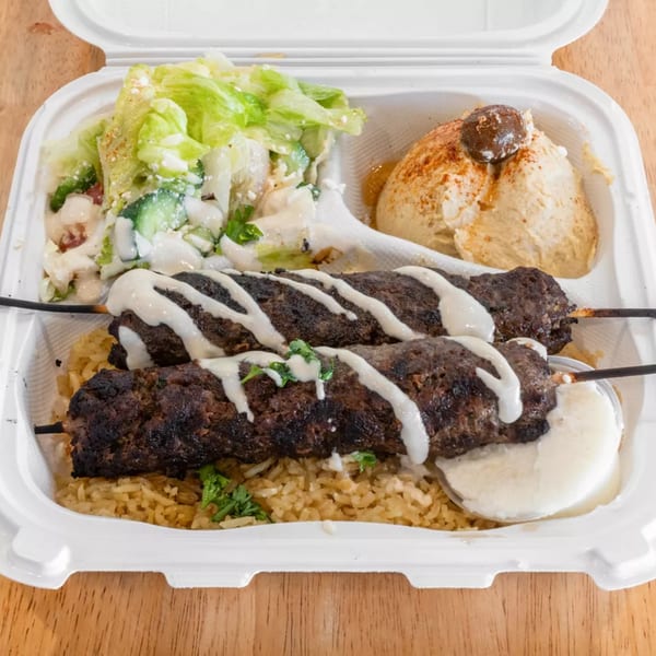 Two grilled meat skewers with white sauce, rice, salad, and hummus in a white takeout container.