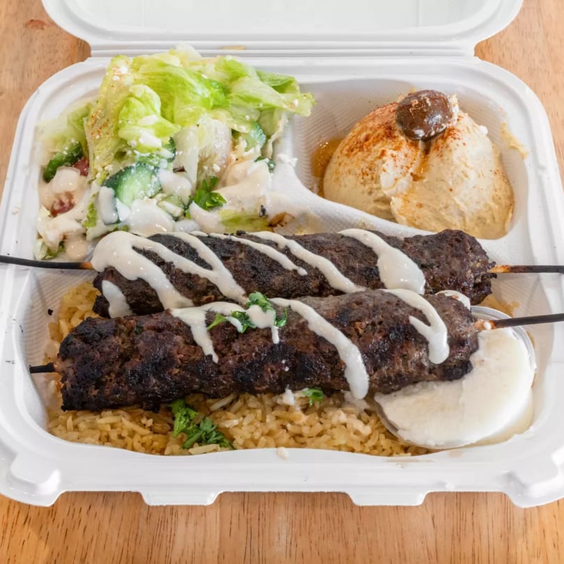Two grilled meat skewers with white sauce, rice, salad, and hummus in a white takeout container.