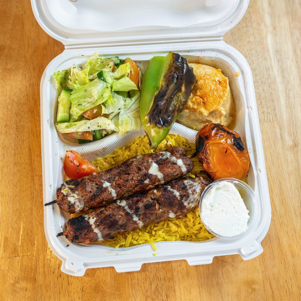Open takeout container with grilled meat skewers, yellow rice, salad, roasted vegetables, and creamy sauce.
