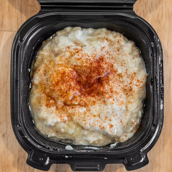 Creamy hummus topped with paprika in a black takeout container on a wooden surface.